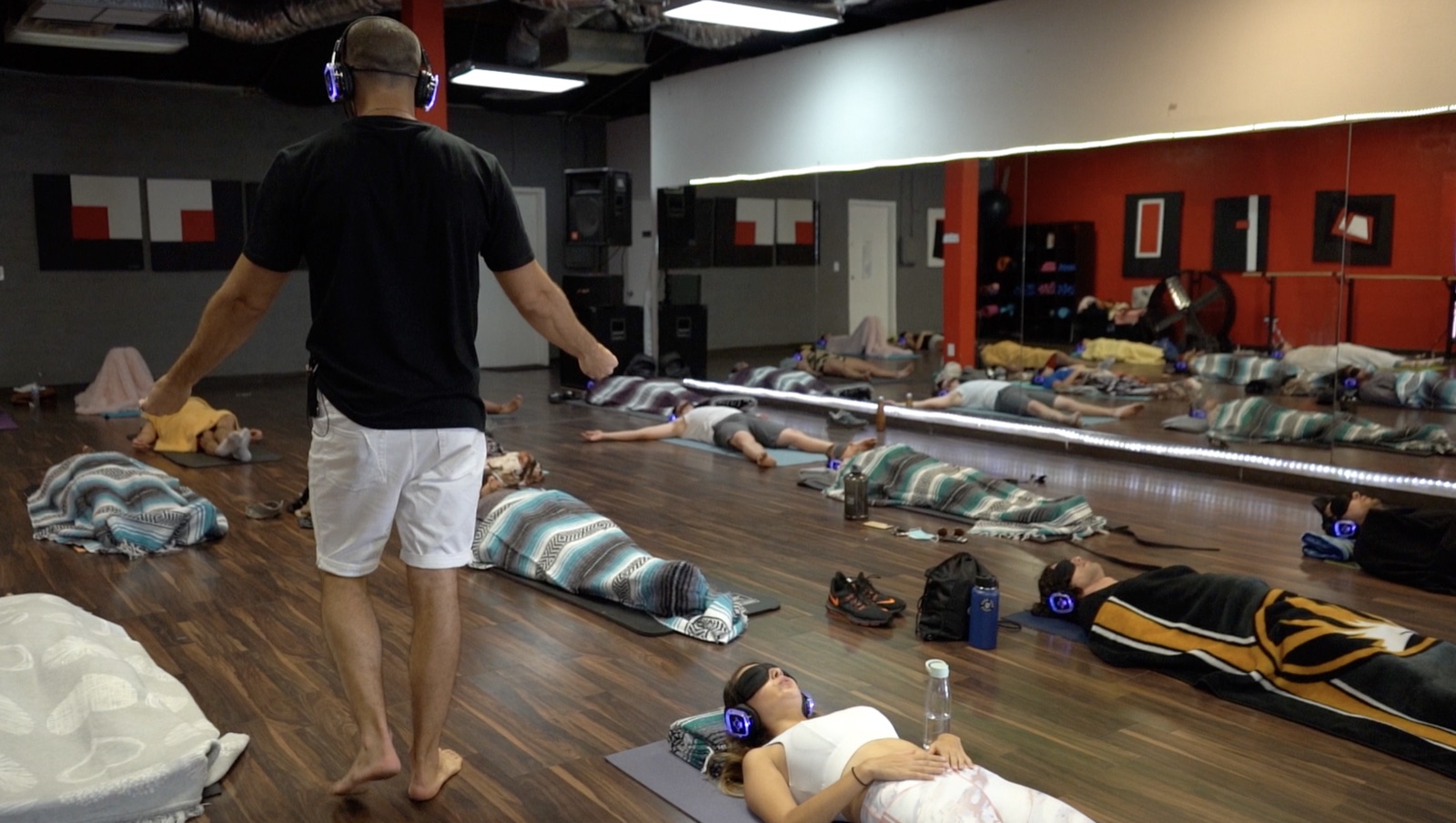 Wide view of a 9D-style headphone group Breathwork session, participants resting on mats with glowing over-ear headphones, each in their own audio bubble even while sharing the same room